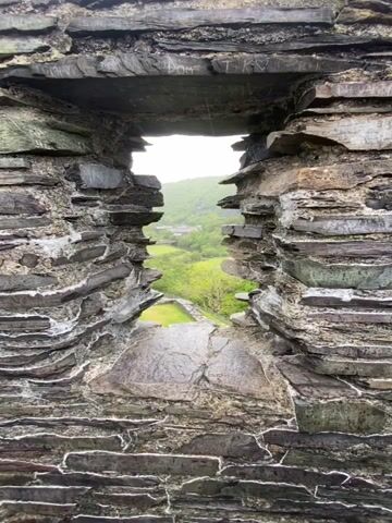 The camera slowly emerges from a stone-walled opening, likely a window or embrasure in an old structure. The opening reveals an expansive view of a green landscape under an overcast sky. Below the opening, the camera focuses on the grass-covered stone foundation of a ruined building, possibly part of the same structure as the window. Beyond the ruins, the view opens to a valley with lush green vegetation, scattered trees, and a body of water in the distance.  A road and possibly some buildings can be seen amidst the trees. The sky is filled with grey clouds, suggesting a damp or rainy day. The walls of the opening are constructed of flat, layered stones.