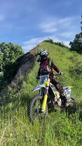 A person in full motocross gear, including a green and black helmet, a red and black long-sleeved shirt with armor, black pants, and boots, sits on a yellow and white dirt bike on a grassy hillside. The person is looking to the left, towards a large body of blue water with small waves visible below the steep incline. Scattered green trees and dark earth are present on the hillside leading down to the water. The sky is blue with a few white clouds.