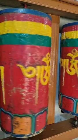 The video shows a row of prayer wheels spinning. The prayer wheels are cylindrical and have colorful bands of yellow, red, and green. They are also decorated with yellow Tibetan script. The prayer wheels are mounted on a wooden frame, and the background appears to be a wall. The video is filmed from a side angle, and the spinning motion of the prayer wheels is evident.
