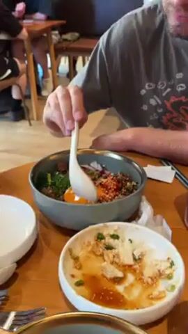 A person is mixing food in a gray bowl with a white spoon. The bowl contains food ingredients including a raw egg yolk, red peppers, chopped vegetables, meat, and herbs. The table is made of wood. The person in the video is wearing a gray t-shirt. A smaller dish with remnants of food sits near the main bowl. The person looks up at the camera and smiles briefly.