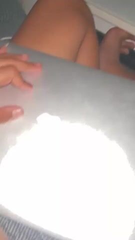 The video shows a person's hands and legs, indicating they are seated or lying down indoors, possibly on a bed or couch, given the striped fabric visible in the background. The scene takes place in what appears to be dim lighting, suggesting evening or night.

The primary object is a silver Apple MacBook laptop. At the beginning, the person's hand rests on the closed laptop, with the iconic Apple logo clearly visible on the lid. The hand then proceeds to open the laptop. As the laptop opens, its keyboard becomes visible, and the screen displays a vibrant blue and white abstract wave-like pattern, characteristic of macOS wallpapers. A small, circular login or user icon is also visible on the screen. The person has a medium to dark skin tone and long fingernails on the visible hand. The location context is Topolchane, Bulgaria. No discernible text is visible on the screen or surrounding objects.