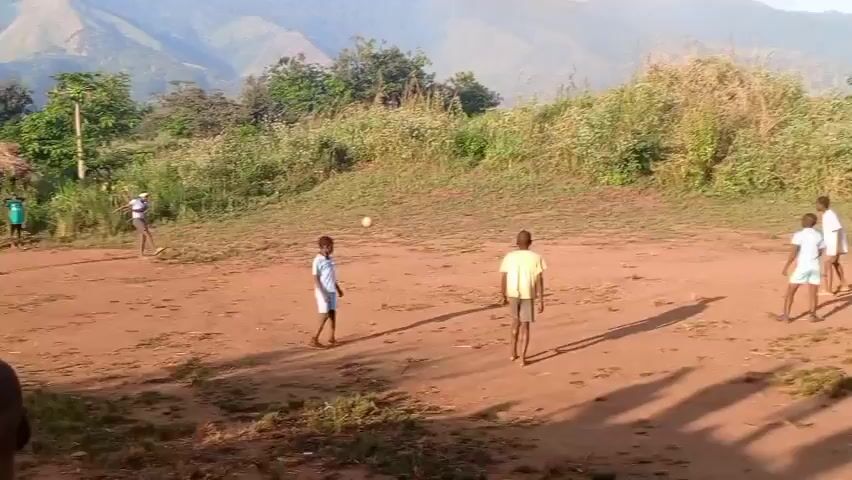 A group of nine children play soccer on a red dirt field. They are wearing a variety of light-colored shirts and shorts, including white, yellow, red, and blue. The soccer ball is kicked around the field. The field is surrounded by grass and bushy vegetation. In the background, there are mountains. The scene is outdoors in daylight.
