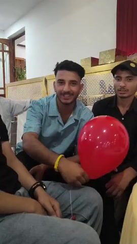 A group of young men are gathered indoors. In the foreground, a person wearing a black shirt faces away from the camera, and another person in a light blue shirt is visible from the side, speaking and gesturing. To the right, a person wearing a black cap is also visible. A large red balloon is prominent in the center of the frame, floating upwards. The background shows a wall with wooden paneling, lighting fixtures on the ceiling, and decorative elements. One man is looking up at the balloon. The interaction appears casual and social, possibly at a party or gathering.