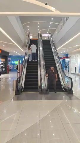 This video shows two escalators in a shopping mall. On the left escalator, two people are ascending. On the right escalator, a person in a dark uniform is descending. The environment is brightly lit with a modern aesthetic.
