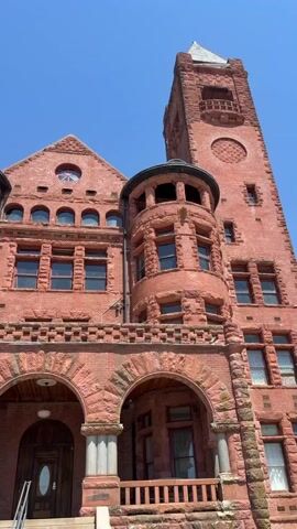 The video shows a wide angle, low-angle shot of a red brick building with a tower. The building has arched doorways and windows. The sky is blue and clear.