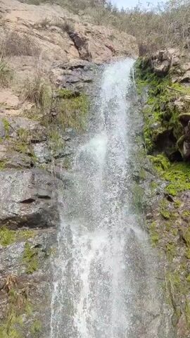 The video displays a close-up view of a cascading waterfall flowing down a rocky, moss-covered cliff face. The water is white and foamy as it plunges into a pool of water at the base of the falls, which is surrounded by rocks. The camera angle is low, looking up at the waterfall.