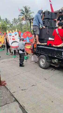 The video shows a street procession featuring people holding large, colorful masks. In the foreground, a man in a light-colored shirt and dark pants walks, appearing to be directing or overseeing the procession. To the right, a person stands on a truck bed, next to a stack of large speakers. Other individuals are visible behind the masks, some of whom are holding flags. The masks are diverse in design, with various facial features and colors. The environment appears to be outdoors, with buildings and trees in the background, suggesting a village or town setting. The overall atmosphere seems festive or ceremonial.