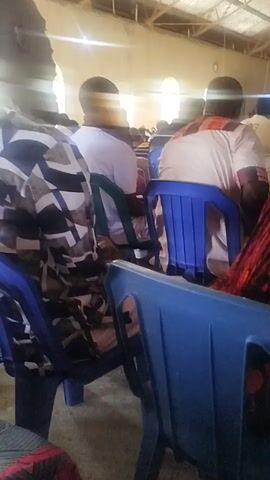 The video shows a gathering of people sitting on blue plastic chairs inside what appears to be a large hall or auditorium. The camera is positioned from behind, focusing on the backs of the attendees. Most of the people are facing forward, with their heads visible. The clothing of the individuals is varied. Several people are wearing headwraps or head coverings. The hall has a high ceiling, and natural light is entering through windows or openings on one side, creating a bright, somewhat diffused light. The building structure seems simple, with a roof made of corrugated metal or a similar material. The video is slightly shaky.