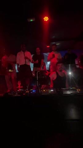 The video depicts a DJ performing in a dimly lit nightclub, surrounded by a small group of people. The DJ, wearing a black tank top, is operating Pioneer DJ equipment, with red lighting emphasizing the controls. Behind him, the group of people are standing, some holding drinks and phones. The background features a screen with a blurry image, further obscured by the red lighting. The setting suggests a lively nightlife atmosphere.