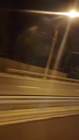 The video shows a nighttime view from a moving vehicle. The camera is pointed out of a window, capturing a blurred roadside scene. A highway or road with yellow and white lines is visible, indicating it's a multi-lane road. There's a guardrail separating the lanes. Streetlights illuminate the scene, creating streaks of light due to the vehicle's movement. In the background, there are dark shapes of trees and some structures, possibly buildings, which are also blurred. The overall impression is one of rapid motion through an urban or suburban area at night.