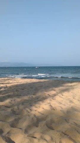 The video shows a beach scene with a boat on the water. The foreground is sandy, with the texture of footprints and shadows.  The midground is the ocean with small waves and a boat. The background is a hazy horizon line with what appears to be mountains in the distance. The sky is clear and blue.