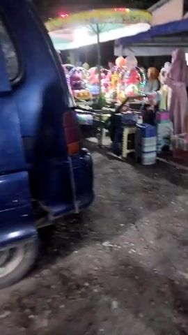 The video, filmed at night in Sambungrejo, Indonesia, captures a lively scene at an outdoor market or fairground. The primary focus is on a small amusement ride, likely for children, which is brightly illuminated with colorful lights. Several children are visible on the ride, suggesting it is in operation.

Surrounding the ride are various stalls and vendors. One prominent stall features an assortment of balloons, with several yellow and green balloons tied together and floating upwards. Another stall displays what appear to be snacks or small items in crates. In the background, a building with a blue and white tarp structure suggests a more permanent market area.

Several people are present, engaged in various activities. A woman in a light purple hijab and dress stands near the balloons, seemingly interacting with the vendor or observing the scene. Other individuals, including men and women, are scattered around the area, some looking towards the ride, others engaged in conversation or browsing stalls. The ground is uneven and appears to be dirt or gravel, indicative of an outdoor, possibly temporary, event space.

The overall atmosphere seems festive and busy, with the glowing lights of the amusement ride and stalls creating a vibrant nighttime ambiance. The presence of families and children suggests a community gathering or local celebration. While no explicit text is visible, the scene is rich with the visual cues of a bustling market event. The time of day is clearly night, and the weather appears clear, allowing for the outdoor festivities.