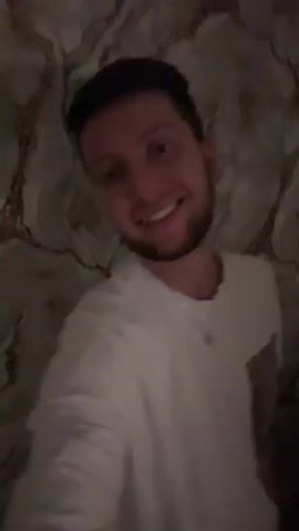The video shows a man with brown hair and a beard wearing a white t-shirt. He is in a room with a patterned wall behind him. The man smiles and makes various hand gestures, including a peace sign and raising his arm. The lighting is dim.