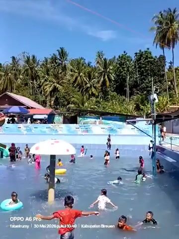 The video showcases a bustling outdoor water park filled with people enjoying the sunny day. The park features a large swimming pool with a tiled bottom, populated with numerous children and adults, some wearing swimwear and others in casual clothing. Many people are wading, swimming, or using inflatable rings. The pool area includes a colorful, spiraling water slide and a mushroom-shaped water feature. Behind the pool, there is a structure with seating, possibly a covered area for relaxation and dining. Beyond that, a lush backdrop of palm trees and other greenery creates a tropical atmosphere. The sky is a clear, bright blue, indicating a warm and pleasant day.