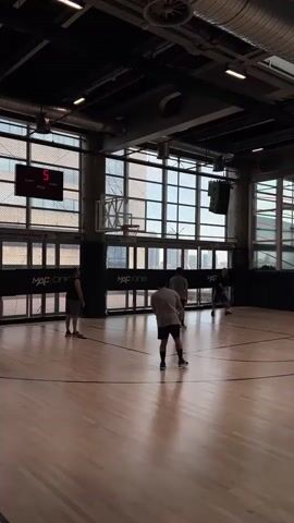 The video shows a basketball court with a wooden floor. There are several people playing basketball. In the background, there is a large window with a view of buildings. A digital scoreboard displays the time and score. The lighting is artificial. The atmosphere appears to be casual and recreational.
