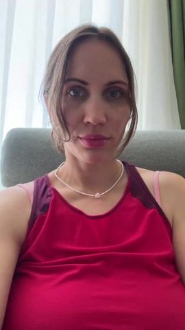 A woman with light brown hair, wearing a red tank top and a pearl necklace, is sitting in a gray chair in front of a window with white curtains. She holds a glass of water containing lemon and cucumber slices, taking a sip. She brings the glass close to the camera, showing its contents.