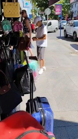 A young boy wearing a white t-shirt, grey shorts, a white baseball cap, and white sneakers stands on a sidewalk next to a market stall selling bags and shoes. The stall is adorned with various bags, including handbags, duffel bags, and luggage. The boy is holding a small object in his hands and appears to be examining it. In the background, a street is visible with cars and pedestrians, suggesting a public outdoor setting. The scene is illuminated by bright sunlight.