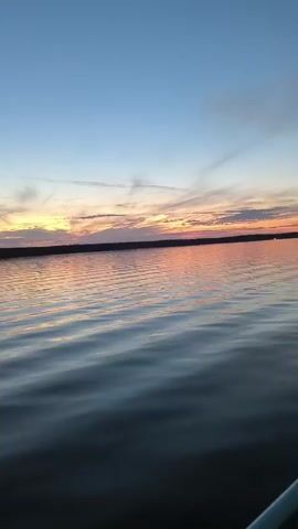 This is a video taken from a boat on a body of water. The water is calm with ripples. In the background, there is a shoreline with trees and a sunset sky. The sky is a gradient of blue, orange, and pink. There are some wispy clouds in the sky. The overall mood of the video is peaceful and serene.