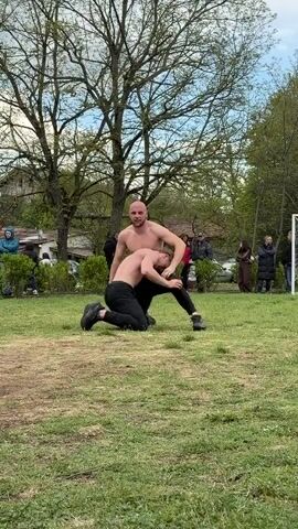 Two shirtless men in black athletic pants are standing on a grassy field. One man is kneeling and touching his head. The other man is standing and looking at the kneeling man. There is a small crowd of people standing behind them. There are trees, a soccer goal, and a few buildings in the background. The sky is overcast.