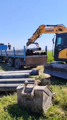 An excavator is unloading pipes from a flatbed truck. The excavator is yellow and black, and the truck is blue. There are several workers on the site, wearing hard hats and safety vests. The scene is outdoors, with a clear blue sky and green grass. The ground is uneven, and there are some trenches in the foreground. The pipes are made of concrete and are being stacked on the ground. The excavator is carefully placing the pipes, and the workers are guiding them into place.