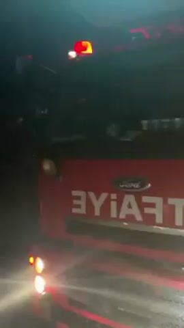 The video shows a red Ford fire truck at night. The truck’s lights are on and the word “itfaiye”, which is Turkish for “fire department”, is visible on the front. The camera pans to the right, showing tree branches and a bright orange glow, suggesting a fire is nearby.