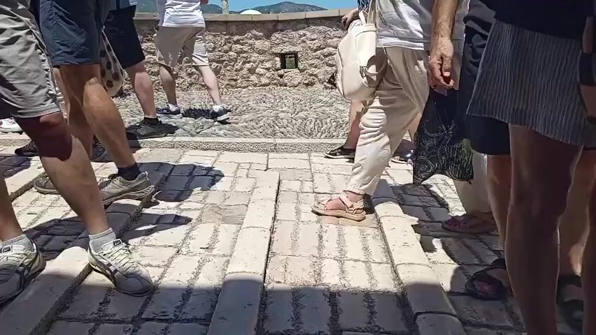 The video displays a low-angle, close-up view of several people's legs and feet walking on a cobblestone path. The path has raised brick sections for walking. The people are dressed in casual summer clothing. The environment appears to be outdoors, with a stone wall in the background and sunlight casting shadows. The individuals are moving in the same general direction, suggesting they are part of a group or a tour. One person on the right is holding a black plastic bag. Another person in the center is holding a white tote bag and walking with a cane. The scene is static, with no camera movement, focusing on the ground level perspective of the pedestrians.