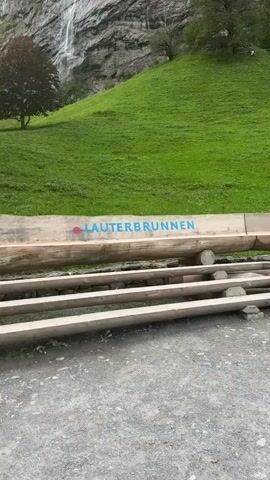 The video shows a scenic view of a waterfall cascading down a rocky cliff face. In the foreground, a wooden bench with the word "LAUTERBRUNNEN" painted in blue letters is visible. The bench is situated on a grassy slope, with a dark-leaved tree to the left and a couple of lighter-leaved trees to the right of the waterfall. The sky is overcast, and the overall atmosphere is serene and natural.