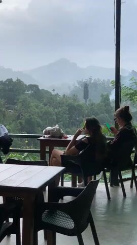 This video shows a view from an outdoor covered seating area overlooking a lush green mountainous landscape shrouded in mist. In the foreground, two wooden tables and chairs are visible. Two women are seated at tables on the right side of the frame. The woman closer to the camera, with long brown hair, is wearing a black dress and is looking down, possibly at her phone or food. The second woman, further back, is wearing a dark top and is also looking at her phone. A person wearing a black t-shirt and shorts is walking from the right towards the tables, carrying a tray with two drinks, one red and one with a white foam top and garnished with lime. Another person, wearing a white hat and a dark jacket, is seated at a table on the left. The environment is overcast and appears to be humid or rainy, contributing to the misty atmosphere of the mountains. A white object, possibly a dog or a stuffed animal, is on the table in front of the first woman. A cell tower is visible amidst the trees in the background.