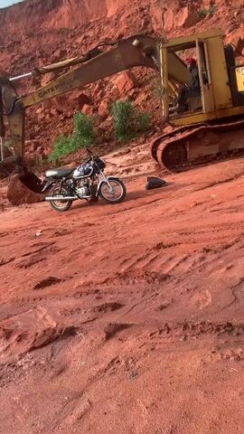 The video shows a construction scene with a yellow Caterpillar excavator on a red dirt path. A motorcycle is parked to the left of the excavator. A person is visible in the driver's seat of the excavator, and another person is standing near the rear of the excavator. The ground is uneven and appears to be a construction site or a recently excavated area. Behind the excavator, there is a steep, red dirt embankment with some greenery at the top. The sky is visible and appears cloudy. The excavator's arm is lowered, and its bucket is on the ground. The motorcycle is upright on its kickstand.