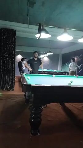 A man stands at a billiard table with a cue stick in his hand. Several other people are visible in the background, also near billiard tables. The environment appears to be an indoor recreational facility with pool tables and overhead lighting. The man in the foreground is dressed in a black t-shirt and blue shorts. He is looking at the billiard balls on the table. The scene is lit by artificial lights.