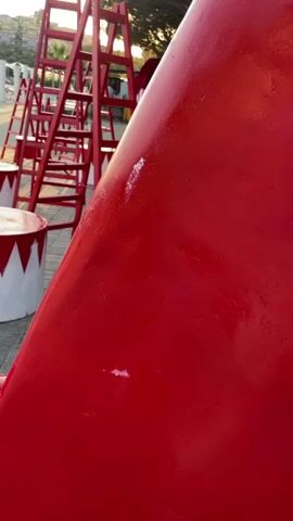 The video shows an outdoor scene decorated with red objects. In the foreground, there is a large, red, bathtub-shaped object. Behind it, there are several red circus-style stools with white tops and red zigzag patterns. Further back, there are multiple red ladders placed upright. To the left, there are also small red tables with mirrors on top. The ground is paved with light-colored interlocking bricks. In the background, there are trees and buildings visible, suggesting a town or city setting near a body of water. The lighting suggests it is either early morning or late afternoon. There are no people visible in the video.