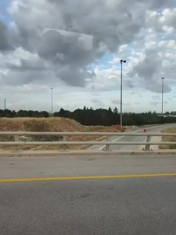 The video shows a scene from a car driving on a bridge. The view is from the passenger side window. A yellow line runs along the right side of the road. A highway with a metal guardrail runs parallel to the road the car is on.  The sky is cloudy. There are several tall lampposts visible. Beyond the highway is vegetation and the outlines of buildings in the distance.  The car's side mirror is visible in the right edge of the frame.
