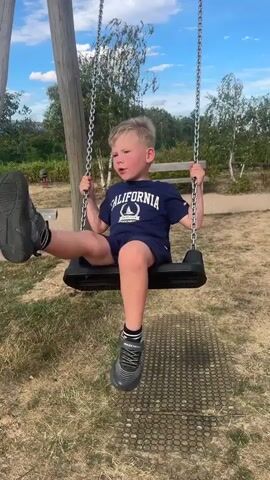 A young Caucasian boy with blond hair is on a playground swing. He is wearing a dark blue t-shirt with "CALIFORNIA" written on it in white letters and dark blue shorts with white trim. He is also wearing dark sneakers with grey accents and white socks. The boy is swinging back and forth, with his legs pumping. The swing set has a wooden post on the left and metal chains attached to a black swing seat. The ground beneath the swing is covered with dry grass and a black rubber mat. In the background, there are trees with green leaves, a wooden bench, and a blue sky with some clouds. The boy appears to be enjoying himself, with a smile on his face.