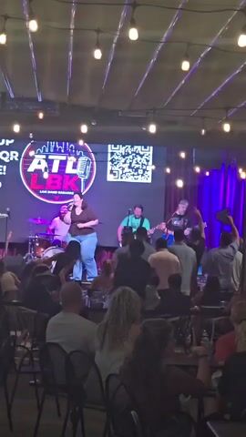 A live band is performing on a stage in front of a large screen displaying "ATL LBK LIVE BAND KARAOKE" and a QR code. The band consists of a drummer, a female singer, and two guitarists. The singer is wearing a brown top and blue jeans. The audience is seated in chairs facing the stage, with some people standing. The venue appears to be a bar or restaurant with hanging lights. The lighting is dim, with stage lights illuminating the performers. The video is shot from the audience's perspective, looking towards the stage.