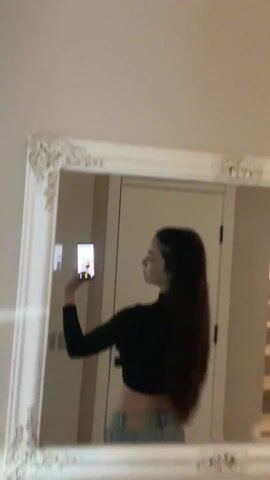 A woman with long dark hair stands in front of a white ornate-framed mirror and takes a picture of herself with her phone. She is wearing a black long-sleeved crop top and light blue jeans. The background is a plain off-white wall and a partially visible light-colored door.