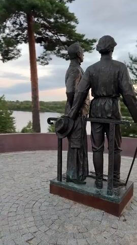 This video shows a bronze statue of two men standing side by side. They are both facing away from the camera and looking towards the left. The man on the left is taller and wears a long coat and boots. He is holding a hat in his left hand and a cane in his right. The man on the right is shorter and wears a suit with a vest and tie. He is pointing his right arm to the left. The statue is on a stone platform, which is surrounded by cobblestones. In the background, there are trees, a body of water, and a cloudy sky. The video is shot from various angles as the camera pans around the statue.