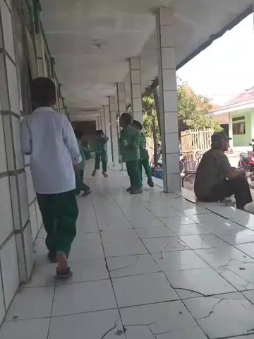 A video shows five children wearing green pants and either white or green tops pushing each other in front of a wall. An older man in a hat and seated position watches them. The scene takes place on a tiled hallway or walkway, possibly at a school or building. The walkway is flanked by pillars and a building with windows on one side, and an open area with trees and bikes on the other.