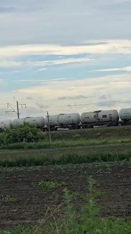 A train comprised of multiple white tank cars is traveling on a track that runs horizontally across the middle of the frame. The train is moving from left to right. The sky above is cloudy with patches of blue. In the foreground, there is a field with dark soil and green plants. The scene is outdoors during the daytime.