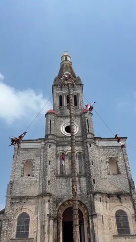 A low-angle shot shows a historical stone church with a tall tower and four people suspended from a pole in front of it. The pole is secured in the ground in front of the church entrance, with the top of the pole extending to the top of the church tower. The individuals appear to be performing some kind of aerial dance, each hanging upside down and slowly spinning on ropes extending from the top of the pole. Two large bird-like figures are attached to the ends of two of the ropes. The sky above is blue with some wispy clouds. The church is built from gray stone, with arched windows and red brick accents. A golden cross sits atop the tower.