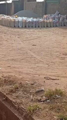 The video depicts a dirt lot with a row of bags stretching into the distance, beside a pile of gravel or sand. The lot also contains a rectangular concrete structure decorated with a mosaic tile pattern and overgrown with weeds.  A concrete drainage ditch runs along the lower left corner of the frame.