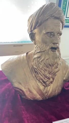 This video shows a golden bust of an elderly man with a long beard and a turban. The bust is placed on a dark red velvet cloth. The background appears to be a room with a window. There is a faint reflection of what might be a picture frame in the upper right corner of the video. The bust is lit from the front, creating shadows on its face. The man's mouth is slightly open, as if he is speaking. The video is shot from a slightly low angle, looking up at the bust. The camera moves slightly from left to right, then zooms in slightly on the bust.