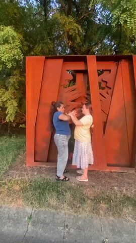 The video shows two young women standing face to face in front of a large, rusty, abstract metal sculpture. The sculpture has cut-out shapes that resemble trees. The woman on the left wears a blue t-shirt and grey pants, while the woman on the right wears a yellow t-shirt and a white floral skirt. They are holding hands and appear to be dancing or posing for a photo. The background consists of green trees and grass, suggesting an outdoor setting. The lighting indicates it might be late afternoon or early evening.