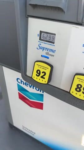 The video shows a close-up view of a gasoline pump at a Chevron station in Kent, USA. The pump is predominantly gray and white with the Chevron logo clearly visible on the side. The digital display at the top of the pump shows the price per gallon for "Supreme" gasoline as $6.299, with "with TECHRON" written below. Below this, a yellow button with the number "92" indicates the minimum octane rating, with "PRESS" printed underneath.

As the video progresses, the display changes. The top digital readout updates to show "This Sale" at $32.29 and "Gallons" at 5.127. The screen in the middle of the pump, which initially displays the Chevron logo and a message about replacing the nozzle, shows a cartoon image of a red car, possibly from the movie "Cars." The surrounding environment suggests it is daytime, with shadows cast on the pump, indicating sunlight. The overall setting is an outdoor gas station, a common scene for refueling vehicles. There are no people visible in the frame, and the focus remains solely on the gas pump and its displays. The activity taking place is the completion of a gasoline purchase, as indicated by the updated sale total and gallons pumped.