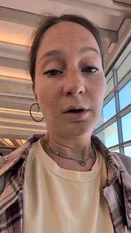 A young woman with long brown hair, freckles, and wearing hoop earrings, a yellow t-shirt, a beaded necklace, and a plaid shirt is filming herself. She appears to be in an airport. She looks up at the ceiling, then down at the camera, then up again. She smiles briefly. In the background, there are large windows, a ceiling with lights, and other people walking.