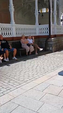 A group of people are sitting on a bench outside a building. The building has a white ornate railing. The people are sitting on a dark colored bench. The person on the far left is a man wearing a black t-shirt and blue shorts. Next to him is a young girl with blonde hair wearing a white top and blue shorts. Next to the girl are two women. The woman to the left is wearing a light brown top and pants. The woman to the right is wearing a white top and brown pants. The woman on the far right is waving. The ground is paved with cobblestones.
