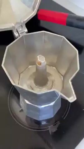 The video shows the top portion of a Moka pot on a black electric cooktop. The Moka pot is made of dull grey metal and the lid is open. Inside the pot is a central metal spout with some brown staining.  A small amount of dark brown coffee grounds are added to the inside of the Moka pot. To the upper right of the pot, slightly out of focus, is a red and black handled tool or object.