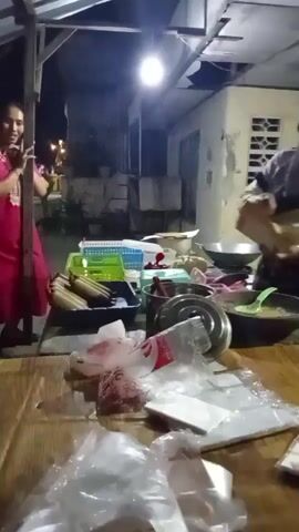 The video shows an outdoor scene at night. A woman wearing a headscarf and an apron is standing at a wooden table, stirring something in a bowl with a spatula. She is holding a piece of paper in her other hand. In the foreground, there are several plastic bags and white packaging on the table. In the background, another woman in a pink dress is standing and appears to be talking or gesturing. There are various containers and objects on a surface behind the table. A light fixture illuminates the scene from above. The environment appears to be an outdoor eating area or market stall.