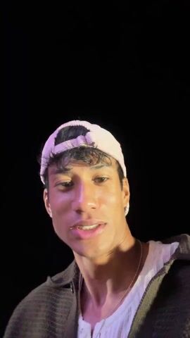 A low-angle selfie video shows a young man on a carnival ride at night. He is wearing a white baseball cap, a grey sweater, and a white t-shirt. To his left, a young woman with long blonde hair is visible, riding in the same seat. They appear to be enjoying the ride, which is brightly lit with various colors. As the ride starts, the camera captures the carnival environment, showing other rides and lights in the background, including one ride labeled “Tornado.” The camera focuses primarily on the young man’s face as he reacts to the ride, looking sometimes at the camera and sometimes around at the environment.