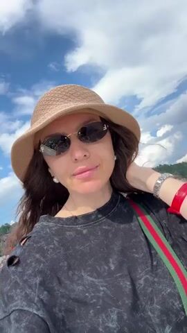 A woman is taking a selfie while on a boat. She is wearing a tan hat, sunglasses, and a black t-shirt with cutouts on the shoulders. She has on a watch and a red bracelet. The boat is moving, and the water is choppy. Mountains are visible in the background, as well as a cloudy sky.