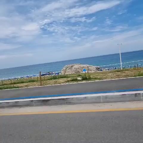 THE VIDEO SHOWS A PERSON DRIVING ALONG A ROAD BESIDE THE OCEAN. ON THE RIGHT SIDE OF THE ROAD, THERE IS A BEACH WITH UMBRELLAS AND A LARGE ROCK. A FEW TREES ARE ALSO VISIBLE ALONG THE COASTLINE. ON THE LEFT SIDE OF THE ROAD, A STREETLIGHT AND A SPEED LIMIT SIGN OF 50 ARE SEEN. THE CAR'S SIDE MIRROR REFLECTS THE DRIVER'S FACE. THE SKY IS BLUE WITH CLOUDS.